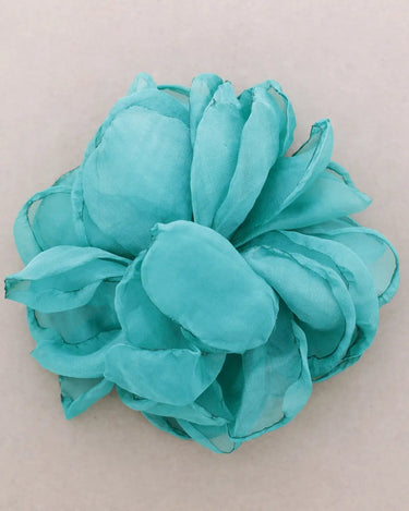 A single artificial flower embellishment in a vibrant turquoise or light blue-green color, made from layered sheer fabric petals, shown on a plain light beige background.