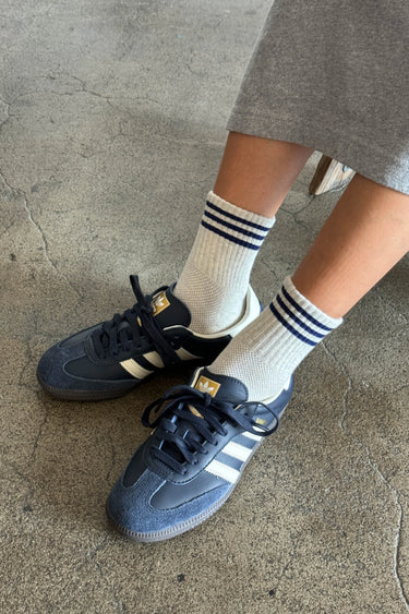 Person wearing navy blue sneakers with white stripes and white socks with blue stripes on a concrete floor.
