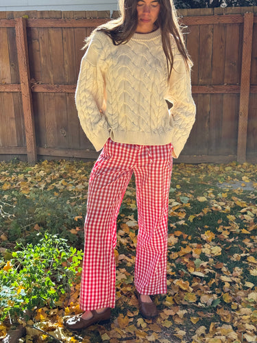 Woman wearing a cream, cable knit sweater and red gingham pants standing on a leaf-covered ground.