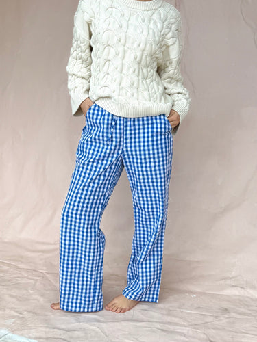 Woman wearing a white cable knit sweater and blue gingham pants on a beige background