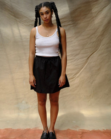 Woman wearing a thin strap tank top and black skirt with braided hair against a beige background.