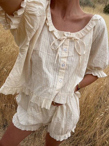 Woman standing in a field wearing a cream eyelet, button-up blouse with a ruffled neckline and matching bloomer-style shorts.