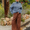 A woman stands outdoors on a sidewalk next to a wooden utility pole, wearing a dusty blue turtleneck sweater and brown wide-leg plaid trousers with brown leather shoes.