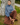 A woman sits on the grass leaning against a large tree trunk, wearing a blue knit mock-neck sweater and brown plaid wide-leg trousers with brown loafers.
