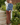A woman stands on outdoor concrete steps beside a white wooden railing, wearing a blue turtleneck sweater and brown plaid trousers, with green plants and a brick wall in the background.
