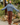 A woman stands outdoors wearing a loose, oversized dusty blue turtleneck sweater and brown wide-leg paid pants, with her arms outstretched. The background features a wooden utility pole, rocks, and green garden plants.