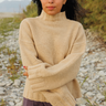 A woman wearing a beige knit mock-neck sweater with ribbed cuffs and hem, standing outdoors on a gravel path with plants and distant hills in the background.