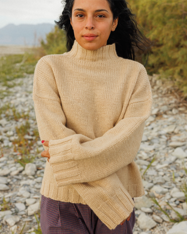 A woman wearing a beige knit mock-neck sweater with ribbed cuffs and hem, standing outdoors on a gravel path with plants and distant hills in the background.
