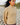 A woman wearing a beige chunky knit mock neck sweater, standing outdoors on a rocky beach with blurred greenery in the background.