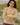 A woman wearing a chunky beige knit mock-neck sweater, standing outdoors with her arms crossed. The background shows a blurred natural landscape with rocks and greenery.