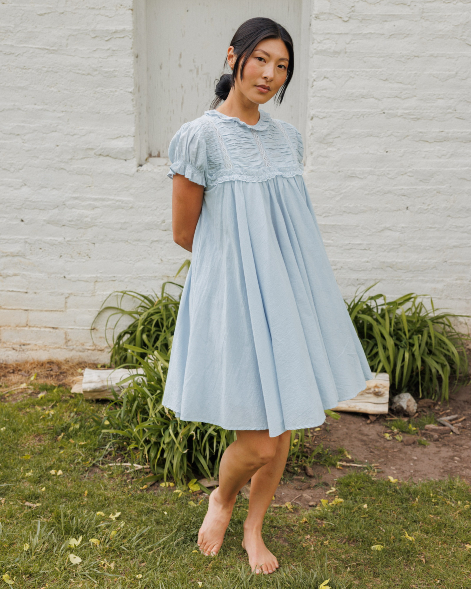 Woman wearing a light blue dress standing outdoors against a white brick wall.