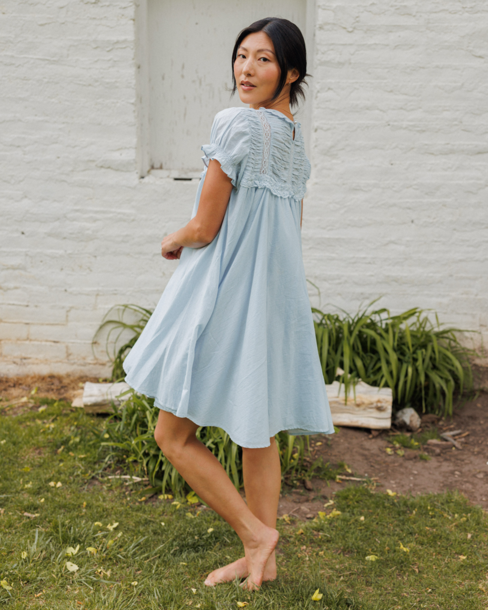 Woman in a light blue dress standing outdoors against a white brick wall.