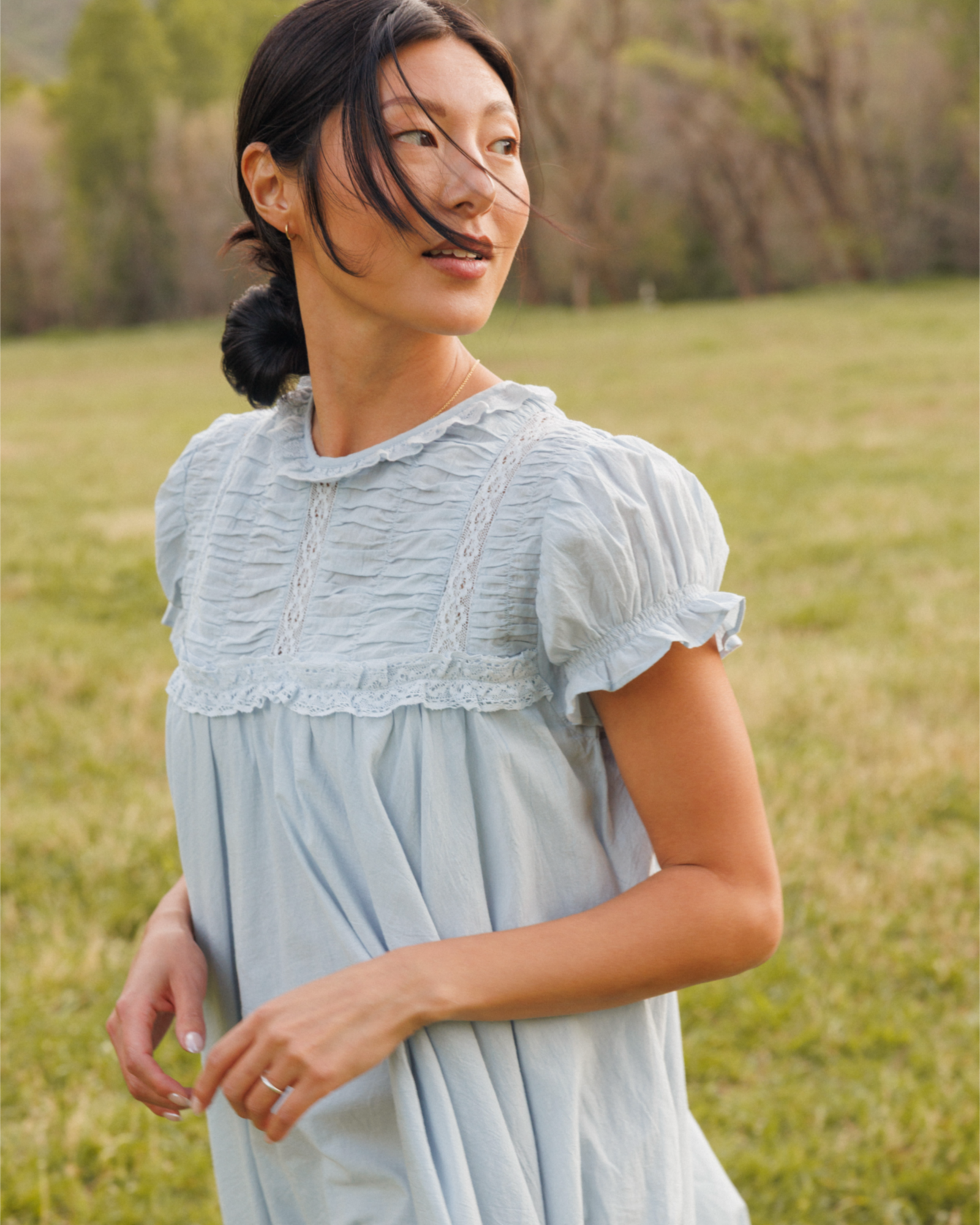 Woman wearing a light blue dress standing in a grassy field with trees in the background