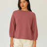 A woman modeling a long-sleeve knit sweater with thin horizontal burgundy and pink stripes, paired with cream-colored pants, against a plain light background.
