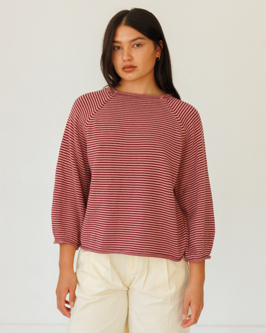A woman modeling a long-sleeve knit sweater with thin horizontal burgundy and pink stripes, paired with cream-colored pants, against a plain light background.