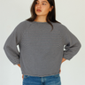 A woman wearing a navy and grey horizontally striped long-sleeve sweater with blue jeans, standing against a plain light gray background.