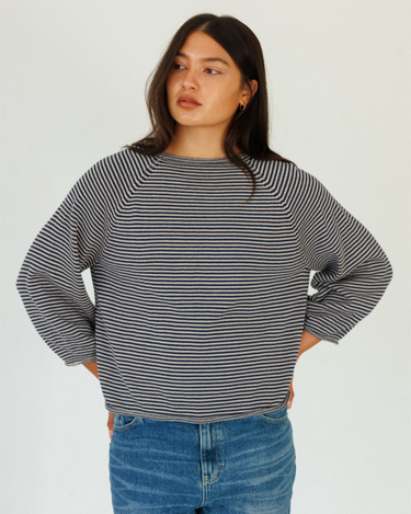A woman wearing a navy and grey horizontally striped long-sleeve sweater with blue jeans, standing against a plain light gray background.