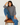A woman sits on a wooden stool wearing a navy blue and grey striped long-sleeve sweater, blue jeans, and brown loafers against a white studio background.