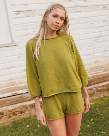 Woman wearing a green knit matching sweater and shorts set standing outdoors with a white wooden structure in the background.
