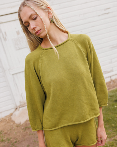 Woman wearing a green knit matching sweater and shorts set standing outdoors with a white wooden structure in the background.