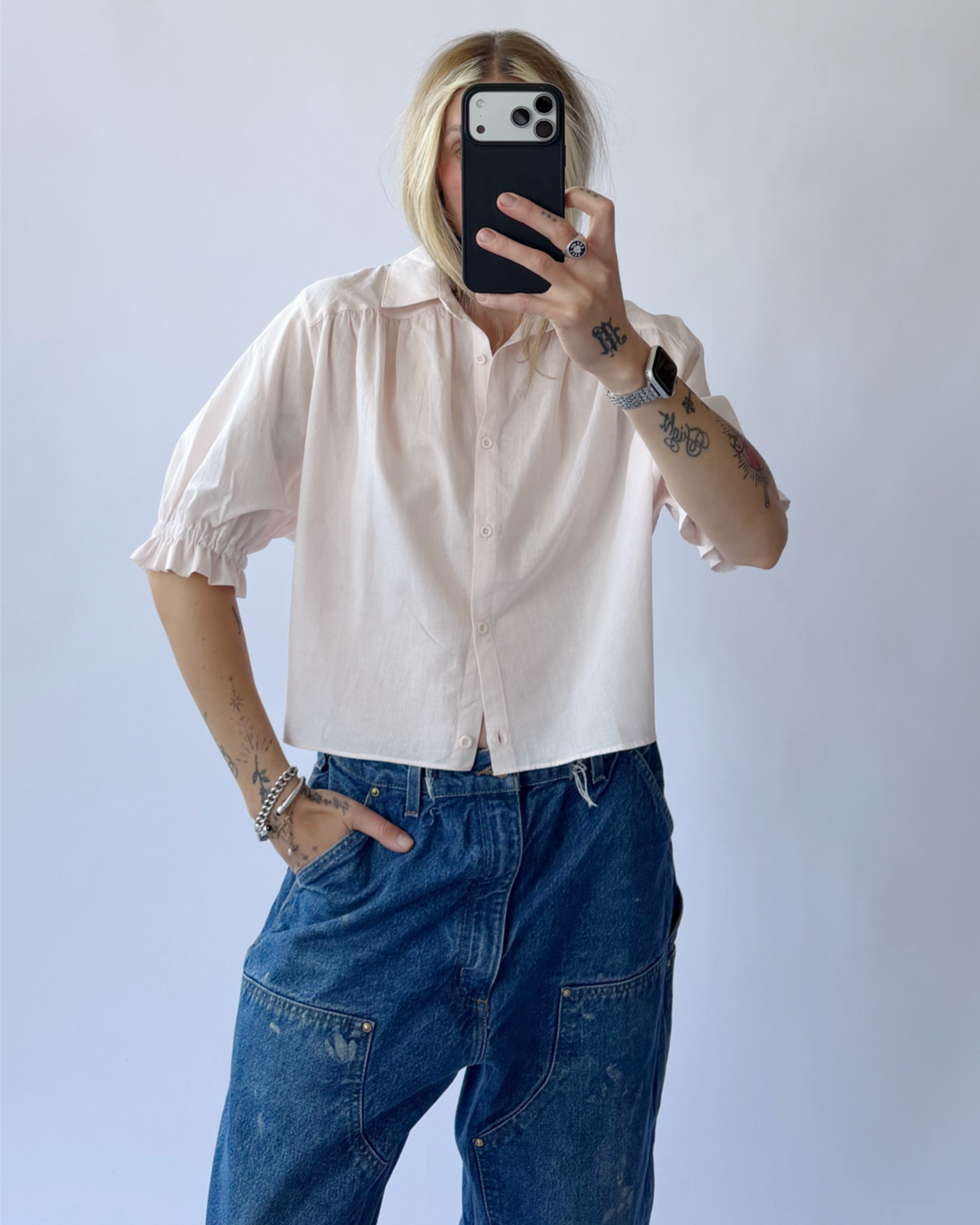 Person wearing a light pink shirt and blue jeans taking a mirror selfie.