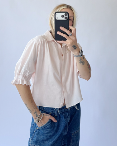 Person wearing a light pink shirt and blue jeans taking a mirror selfie.