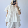 Person wearing a white outfit with ruffled sleeves against a plain background