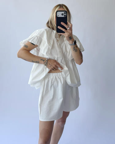 Person wearing a white outfit with ruffled sleeves against a plain background
