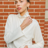 A woman wearing a cream-colored long-sleeve mock neck top with a buttoned collar detail, posed against a red brick wall background.