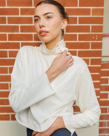 A woman wearing a cream-colored long-sleeve mock neck top with a buttoned collar detail, posed against a red brick wall background.