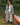 A woman stands in shallow water by a lake wearing a light gray cropped cardigan over a white top and loose taupe wide-leg pants, with a red canoe visible at the right edge.