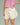 Close-up of a woman wearing a pink top and cream shorts on a green background.