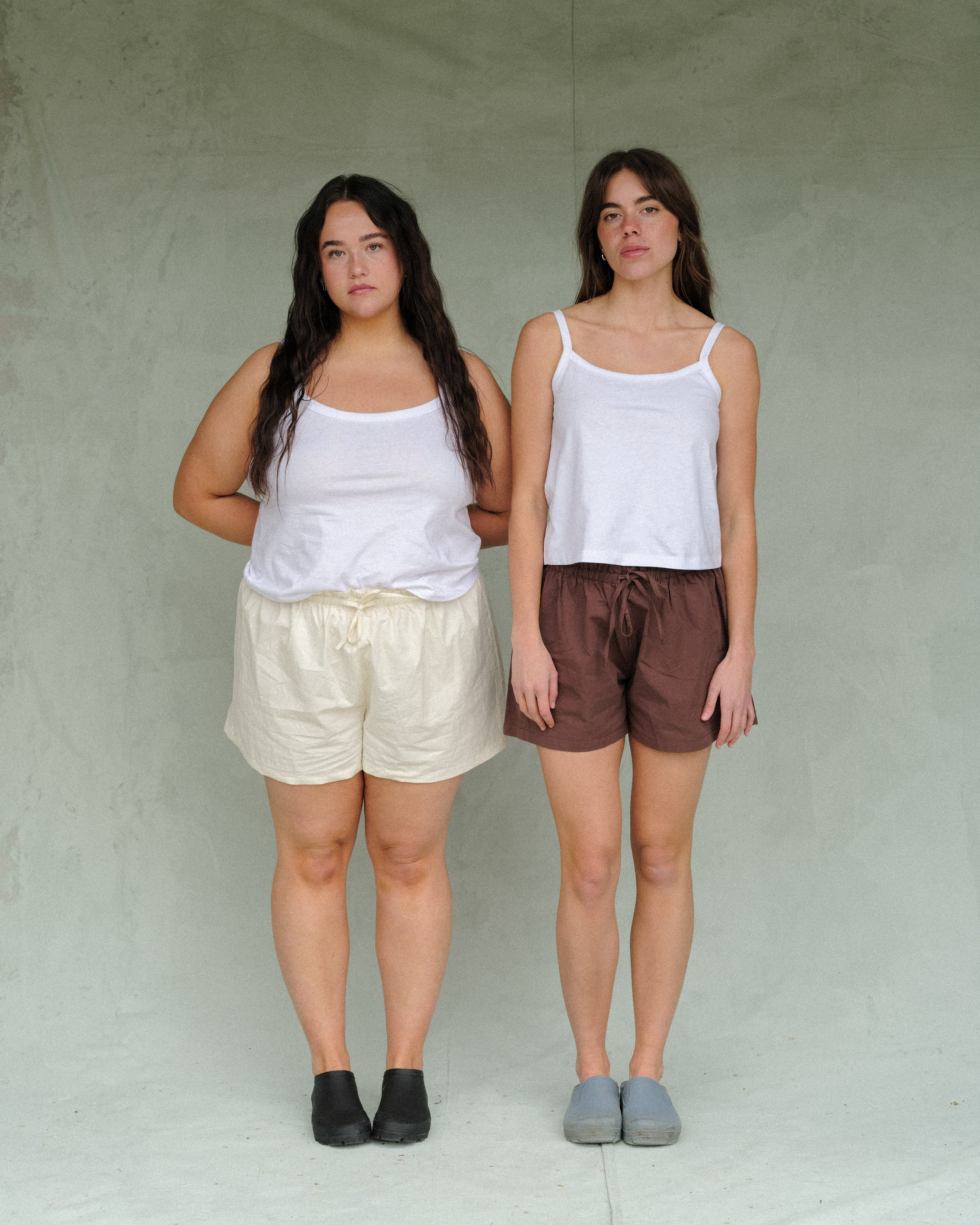 Two women standing next to each other on a green background. One woman is wearing a white tank top with cream shorts, the other woman is wearing a white tank top with brown shorts.