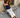 Woman sitting on a stone step wearing a navy blue tank top and cream pants, with a brick wall and wooden door in the background.