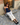 Woman sitting on a stone step wearing a navy blue tank top and cream pants, with a brick wall and wooden door in the background.