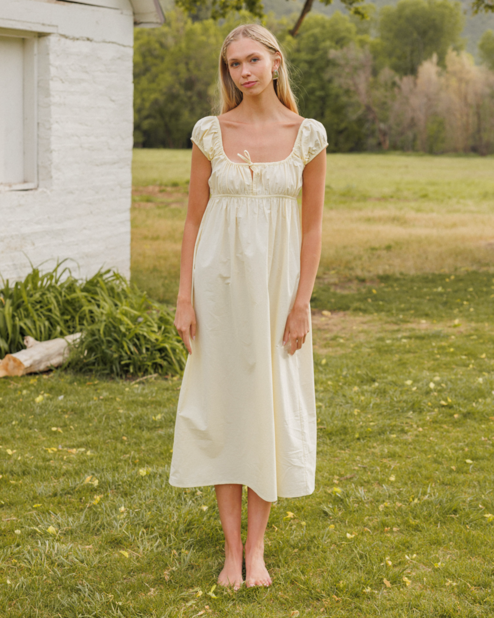 Woman in a yellow dress standing in a grassy field with a white building and trees in the background.