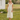 Woman in a yellow dress standing in a grassy field with a white building and trees in the background.