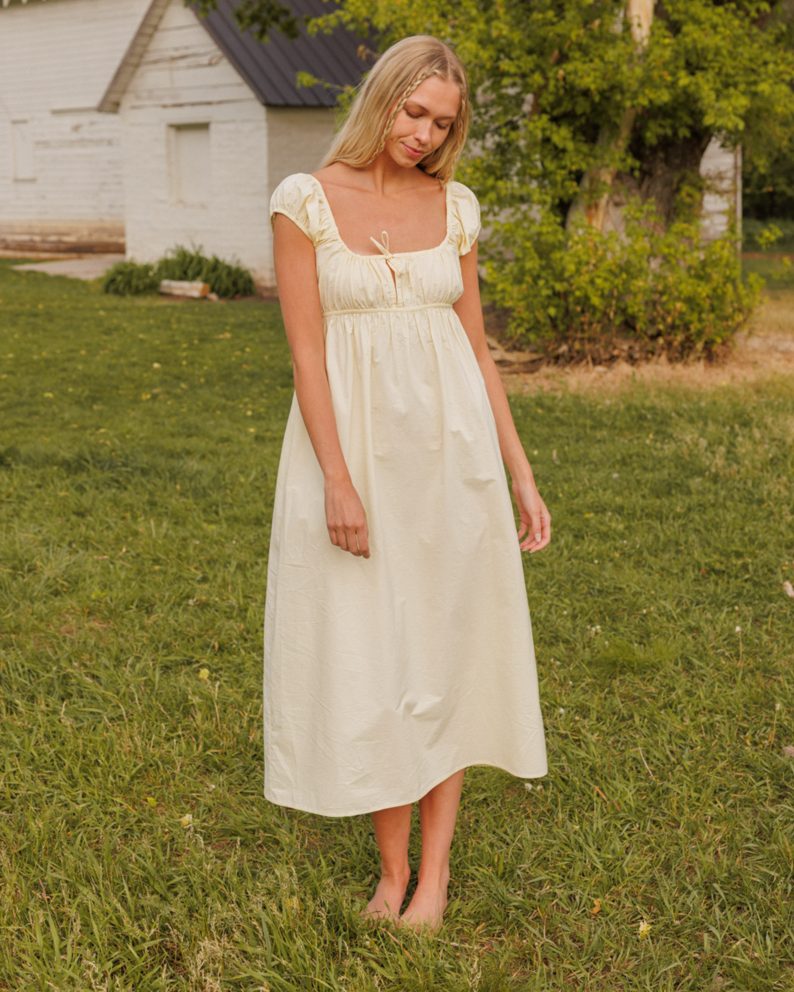 Woman in a yellow dress standing in a grassy field with a rustic building in the background