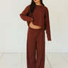 A woman modeling a matching dark red lounge set consisting of a cropped long-sleeve top and wide-leg pants, standing against a white wall and light wood floor.