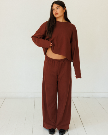 A woman modeling a matching dark red lounge set consisting of a cropped long-sleeve top and wide-leg pants, standing against a white wall and light wood floor.