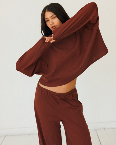 A woman modeling a matching dark red lounge set consisting of a cropped long-sleeve top and wide-leg pants, posed against a clean white studio background.