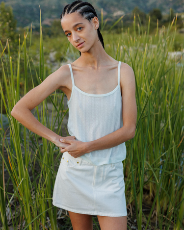 Woman standing in front of a field with long grass, wearing a pointelle tank top and white denim skirt.