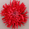 A bright red fabric flower with layered, spiky petals arranged in a round daisy-like shape, photographed from above on a plain light gray background.