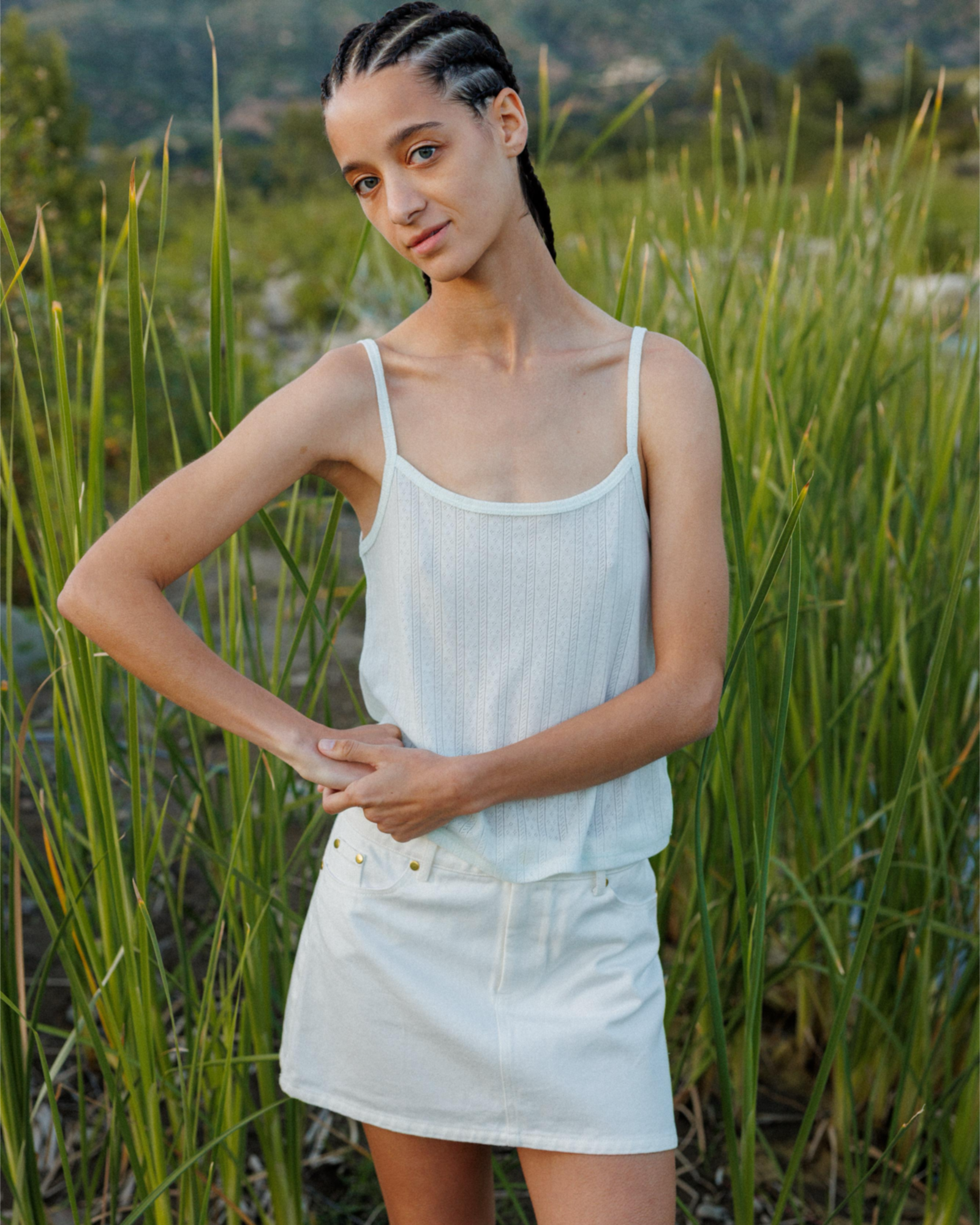 Woman standing in front of a field with long grass, wearing a pointelle tank top and white denim skirt.