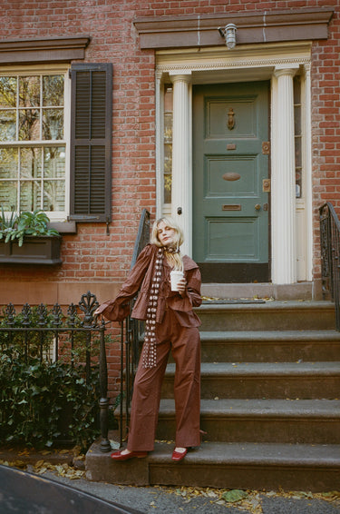 A woman standing in front of a house wearing a brown top with a sailor-style collar and matching brown pants with an elasticated waistband and cuffed hems. The outfit features drawstrings and the top is tied at the front.
