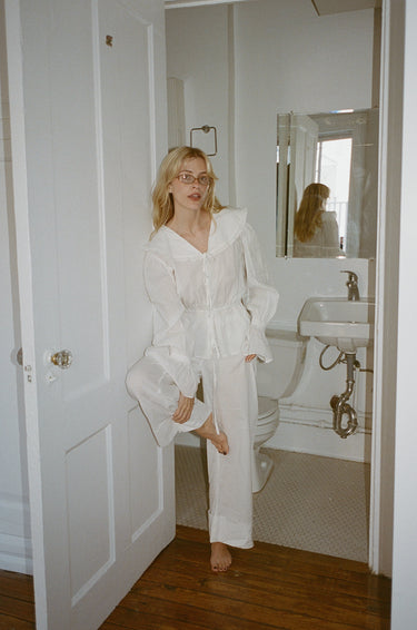 A woman standing in the doorway of a bathroom wearing a white top with a sailor-style collar and matching white pants with an elasticated waistband and cuffed hems. The outfit features drawstrings and the top is tied at the front.