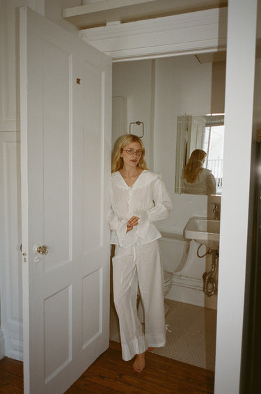 A woman standing in the doorway of a bathroom wearing a white top with a sailor-style collar and matching white pants with an elasticated waistband and cuffed hems. The outfit features drawstrings and the top is tied at the front.