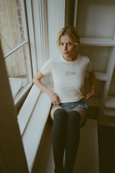 Woman sitting near a window wearing a cream t-shirt with brown stamp-style graphics.