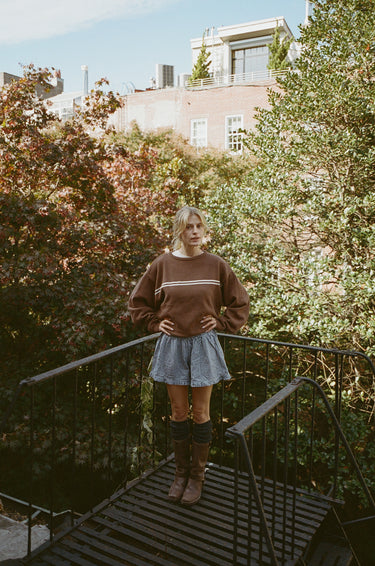 Woman standing on a black staircase outdoors, wearing a brown crewneck sweater with two white stripes and blue plaid boxer-style shorts.