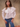 A woman wearing a white blouse with a large ruffled collar and blue jeans on a tan fabric background.